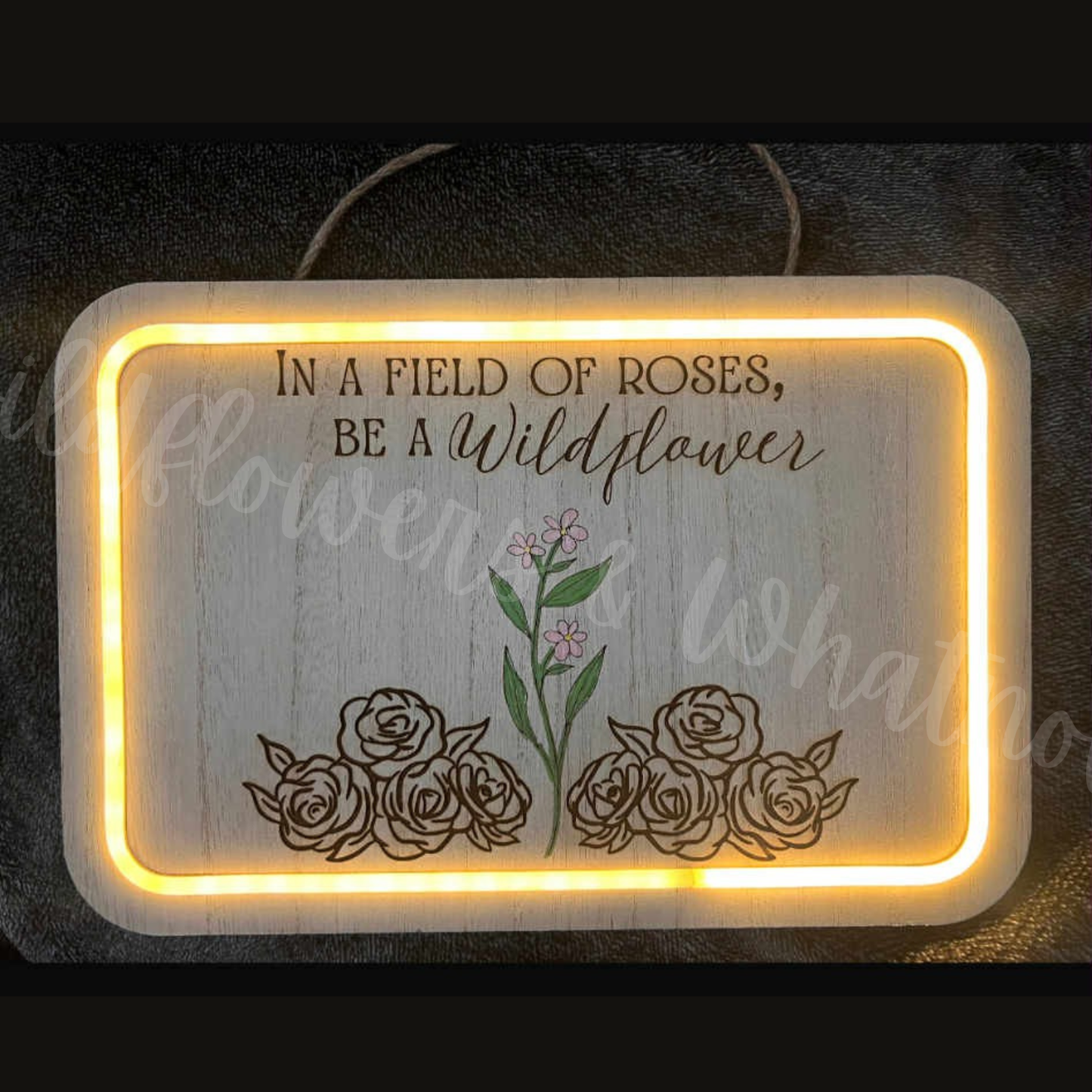 Decorative sign with floral design and text 'In a field of roses, be a wildflower' on a dark background.